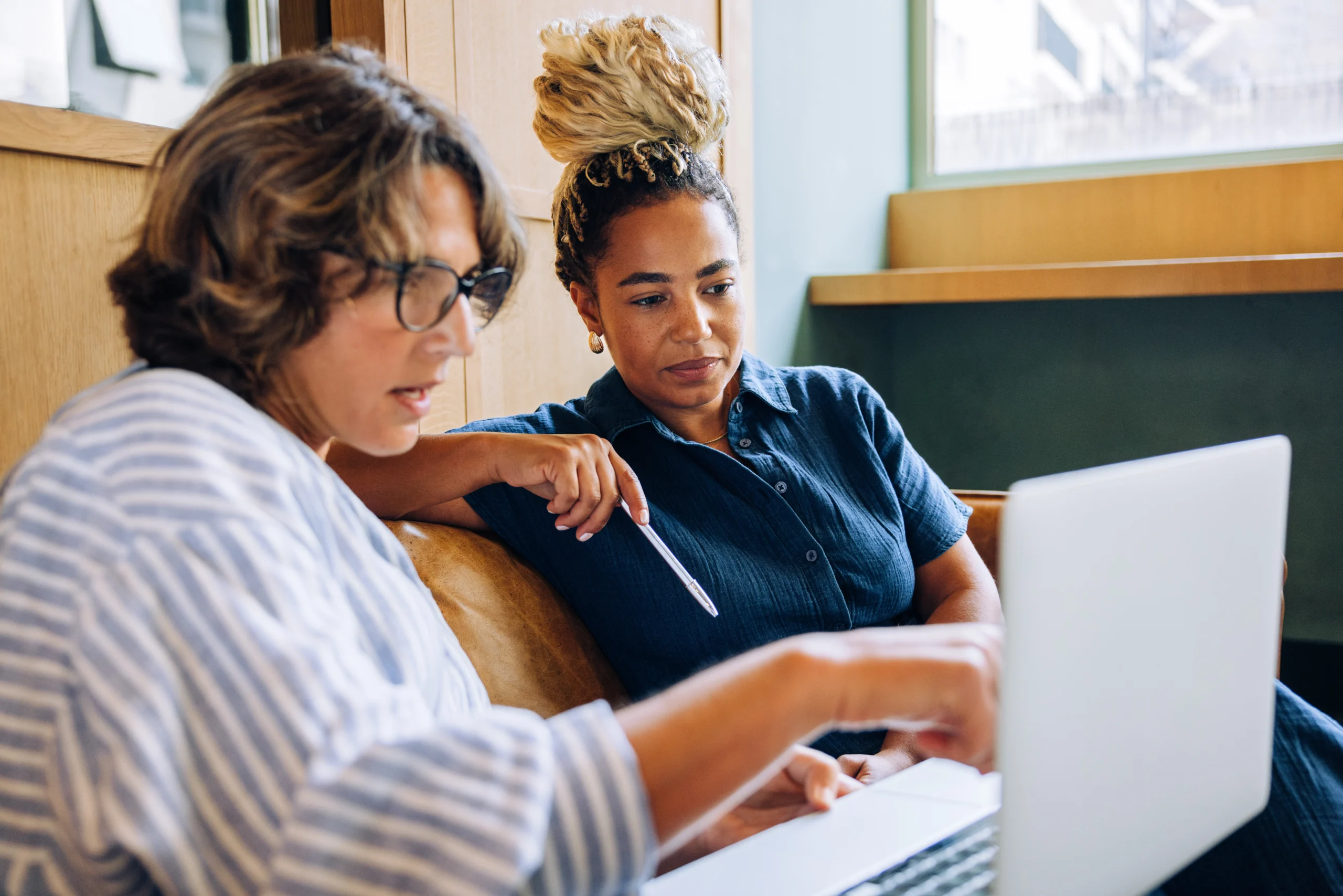 Two women discussing work together while looking at a laptop screen Two businesswomen sitting indoors, engaged in a focused conversation, examining a laptop screen nearby.