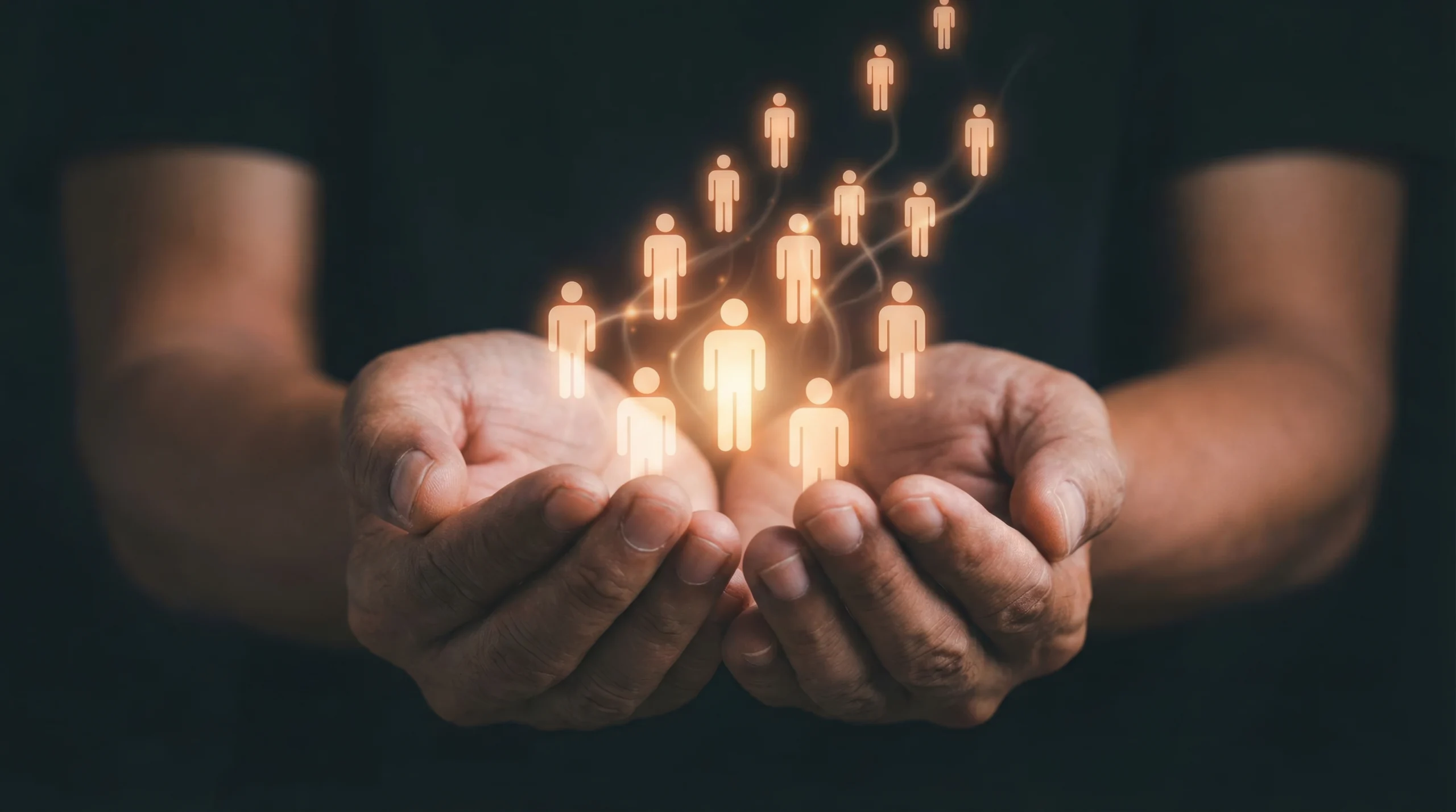 cupped hands cupped hands holding glowing orange people icons floating upward on dark background.