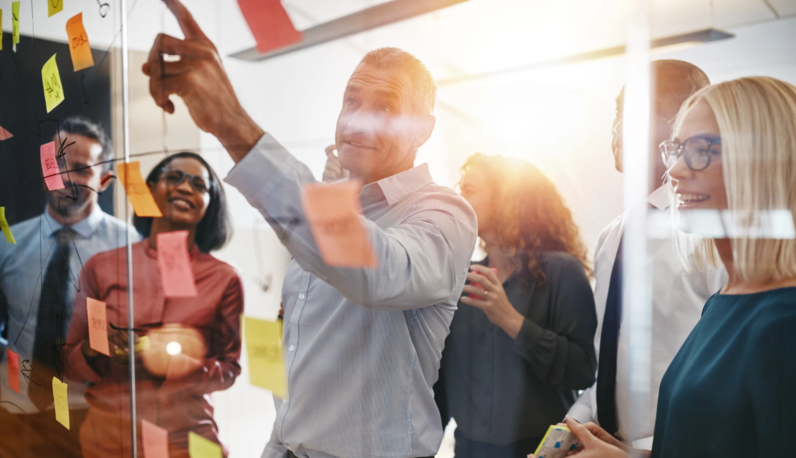 Businesspeople brainstorming together with sticky notes in an of Smiling mature manager pointing at sticky notes on a glass wall while brainstorming with his diverse team in a modern office